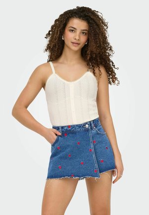 Ung kvinde med krøllet hår iført en hvid strikket ærmeløs top og en blå denim miniskørt med små røde hjerte-mønstre.