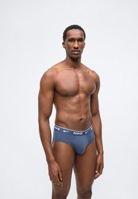 Μυώδης άνδρας που φορά μπλε εσώρουχα Nike, στέκεται μπροστά σε απλό φωτεινό φόντο, κοιτάζοντας προς τα εμπρός με ουδέτερη έκφραση.