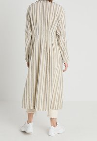 Pruhované béžové šaty s dlouhými rukávy, zúženým pasem a splývavou sukní; mají vertikální modré pruhy, texturovanou látku a uvolněný střih.