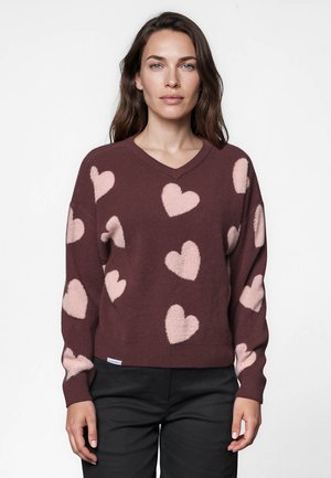 Femme portant un pull marron à col en V avec des motifs de cœurs roses et un pantalon noir, debout devant un fond blanc uni.
