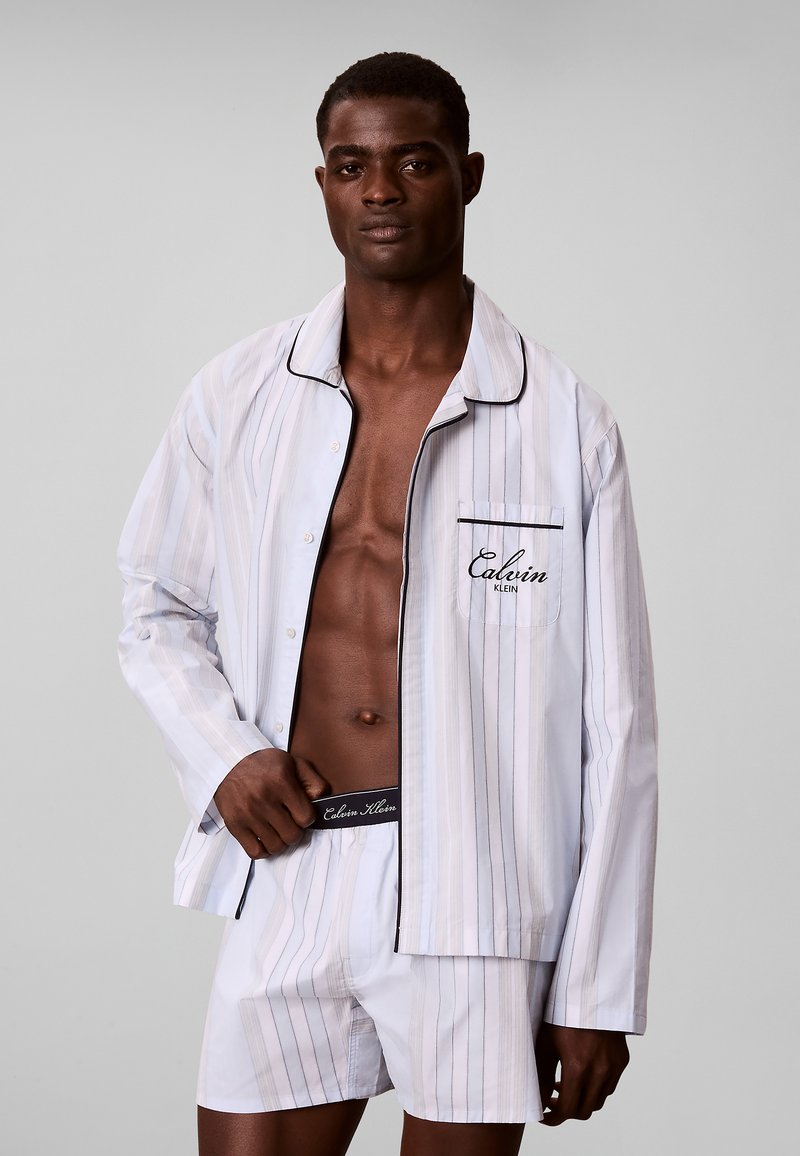 Man wearing white striped Calvin Klein pajamas with open shirt showing bare chest against a plain gray background.