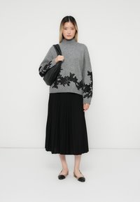 Femme portant un pull col roulé gris à motifs floraux noirs, une jupe midi plissée noire, des ballerines bicolores et tenant un sac à dos noir.