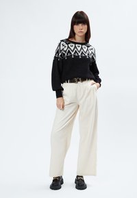 Schwarzer Pullover mit weißen geometrischen Mustern, cremefarbene Hose mit Falten und schwarze Schuhe mit goldener Kettenverzierung.