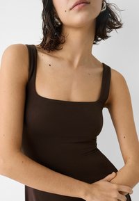 Brun linne med fyrkantig halsringning och breda axelband, gjort av ett mjukt, elastiskt material. Närbild som framhäver hudens textur.