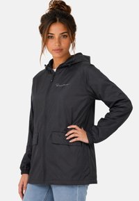 Jeune femme aux cheveux attachés en chignon, vêtue d'une veste à capuche noire avec poches avant et un jean bleu clair sur un fond uni.