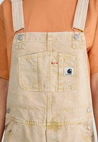 Béžové deninové overaly majú nastaviteľné ramienka so striebornými komponentmi, veľkú prednú vrecko a nášivku s logom Carhartt. Pod nimi je oranžová košeľa.
