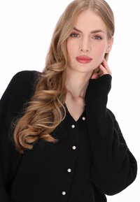 Fekete kardigán V-nyakú, hosszú ujjú, bordázott mintázattal, gyöngyszerű gombokkal az elülső részen. Simított és testhezálló szabás.