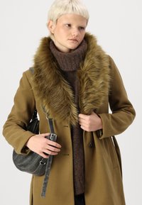Brun frakke med en falsk pelskrave, parret med en mørk struktureret sweater. Modellen holder en sort halvmåneformet taske med en rem.