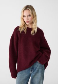 Pull en tricot bordeaux avec une coupe décontractée, un col rond et des manches longues. Porté par-dessus un jean bleu clair, avec une texture douce et moelleuse.