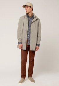 Hellgraue wasserdichte Jacke mit hohem Kragen, blauer Blümchenmuster-Hemd, braune Hose und beiger Cap. Die Schuhe sind leicht und mit einer strukturierten Oberfläche.