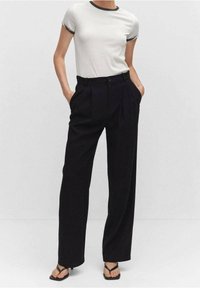 Zwarte hoge taille broek met plooien, gecombineerd met een witte blouse met korte mouwen en zwarte afwerking; model staat met handen in de zakken.