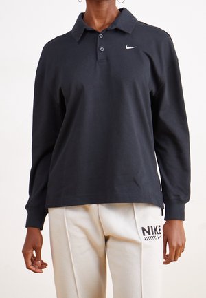 Person iført en sort langærmet Nike polo og off-white Nike joggingbukser med synlige logoer på begge beklædningsgenstande.