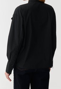 Schwarz Langarmshirt mit lockerer Passform, aus glattem Stoff. Verfügt über Manschetten mit Knöpfen und einen einfachen Kragen, ohne sichtbare Muster.