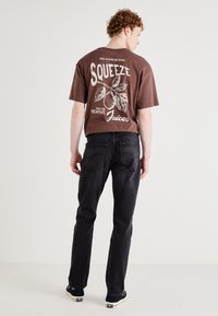 Brown graphic t-shirt with white citrus design, paired with dark denim jeans. Model shown from behind, wearing black shoes with white soles.