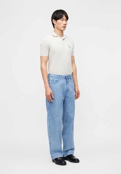 Νεαρός άνδρας στέκεται, φορώντας λευκό πόλο με κοντά μανίκια, ανοιχτό μπλε τζιν παντελόνι με φαρδιά μπατζάκια και μαύρα παπούτσια, μπροστά σε απλό λευκό φόντο.