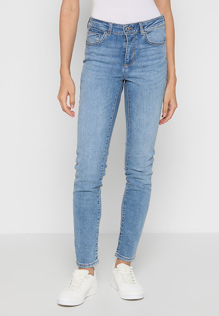 Jean skinny bleu clair avec taille haute, passants de ceinture, poches avant et couture contrastée, associés à des baskets blanches.