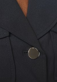 Blazer marine à la texture lisse, avec un gros bouton rond brillant et des revers prononcés, détaillé de surpiqures contrastantes.