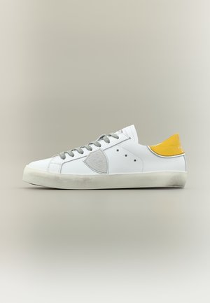 Baskets basses blanches avec lacets gris, patch latéral gris, languette de talon jaune et semelle blanche légèrement usée sur un fond gris clair.