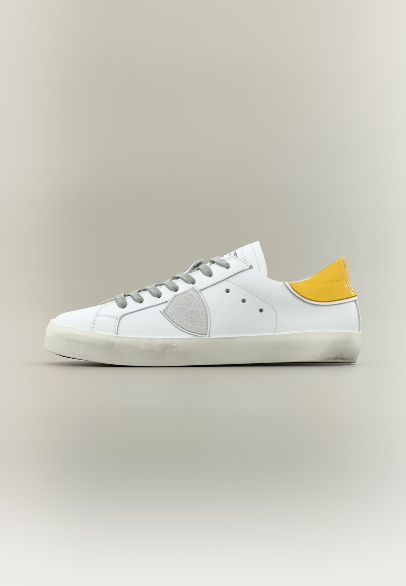 Baskets basses blanches avec lacets gris, patch latéral gris, languette de talon jaune et semelle blanche légèrement usée sur un fond gris clair.