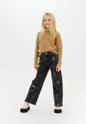 Beige gestrickter Pullover mit verzierten Perlen; schwarze Jeans mit goldenen himmlischen Mustern; schwarze Plateauschuhe. Einfaches, lässiges Outfit.