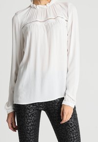 Hvid langærmet bluse med rynket halsudskæring, blonde detaljer over brystet og en flydende silhuet. Parret med sorte teksturerede leggings.
