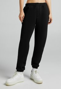 Person i svarta joggingsbyxor med elastisk midja och vita sneakers står mot en enkel grå bakgrund.