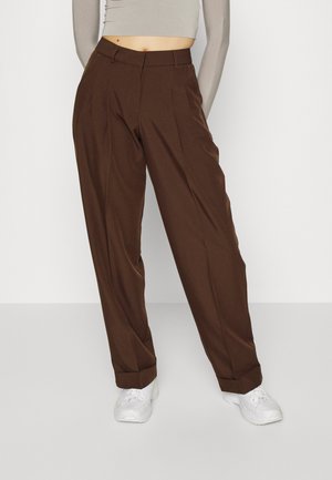 Femme portant un pantalon marron taille haute à jambes larges avec revers et des baskets blanches, associé à un top beige court à manches longues.