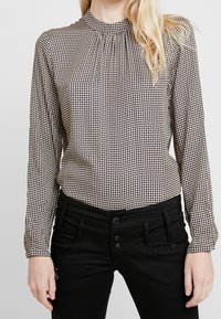 Blus med långa ärmar i ett mönstrat tyg med ett brunt och krämfärgat rutmönster, med en veckad halsringning och svarta byxor med knappar.