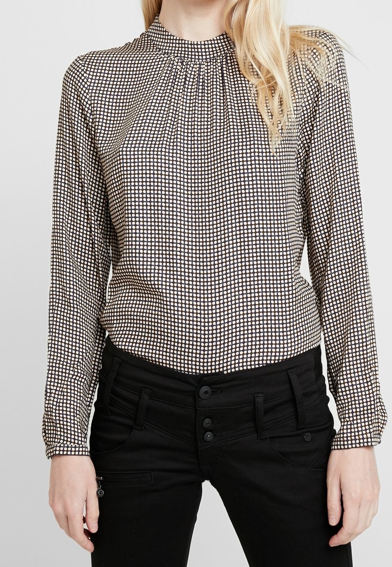 Blus med långa ärmar i ett mönstrat tyg med ett brunt och krämfärgat rutmönster, med en veckad halsringning och svarta byxor med knappar.