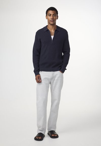 Marinefarbener Strickpullover mit einem halben Reißverschluss am Kragen, graue Jeans und schwarze Badesandalen. Das Modell steht aufrecht vor einem schlichten hellen Hintergrund.