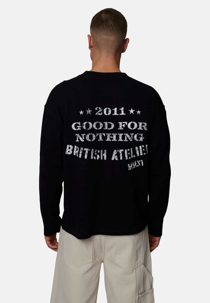 Homme de dos portant un haut noir à manches longues avec le texte blanc « 2011 Good for Nothing British Athletic MMXI » et un pantalon beige.