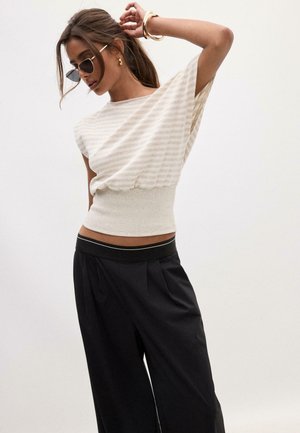 Frau mit Sonnenbrille, ärmellosem beige gestreiftem Oberteil mit tailliertem Schnitt und lockeren schwarzen Hosen, posiert mit einem Arm hinter dem Kopf erhoben.