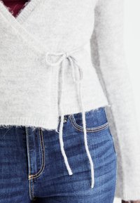 Hellgrauer, weicher Strickwickelsweater mit einer Bindeschließe in der Taille. Er hat lange Ärmel und einen leicht verkürzten Saum, kombiniert mit dunkelblauen Jeans.