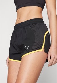Pantalones cortos deportivos negros con ribete amarillo, que cuentan con una cinturilla elástica y aberturas laterales para mayor movilidad. Logotipo visible en el lado izquierdo.