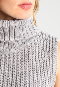 Grå stickad ärmlös polotröja med ruggad textur, som har en hög, vikt krage och en åtsittande silhuett.