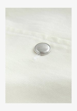 Zilverkleurige ronde knop bevestigd aan wit textiel met zichtbare stiksels.