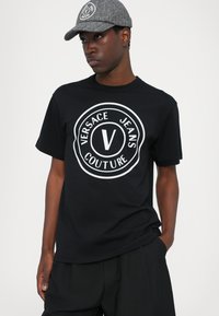Svart bomull t-skjorte med hvit sirkulær logo "VERSACE JEANS COUTURE" på forsiden. Grå caps med lignende logo. Teksturert stoff på capsen.