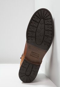 Chaussure en daim marron avec une semelle en caoutchouc noir texturé, présentant des rainures de bandeau à motifs et un petit logo "REDSKINS" sur le talon.