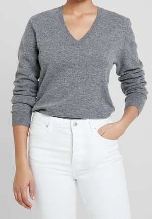 Osoba ubrana w szary sweter z długim rękawem i dekoltem w serek oraz białe wysokie jeansy, z jedną ręką w kieszeni, stoi na jednolitym tle.