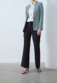 Blazer bleu clair à revers cranté, blouse blanche avec un nœud papillon, et pantalon noir évasé. Des escarpins noirs à brides complètent la tenue.
