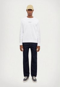 Homem com boné bege, camisa branca de manga longa Michael Kors, jeans escuros e tênis cinzas, de pé contra um fundo branco liso.