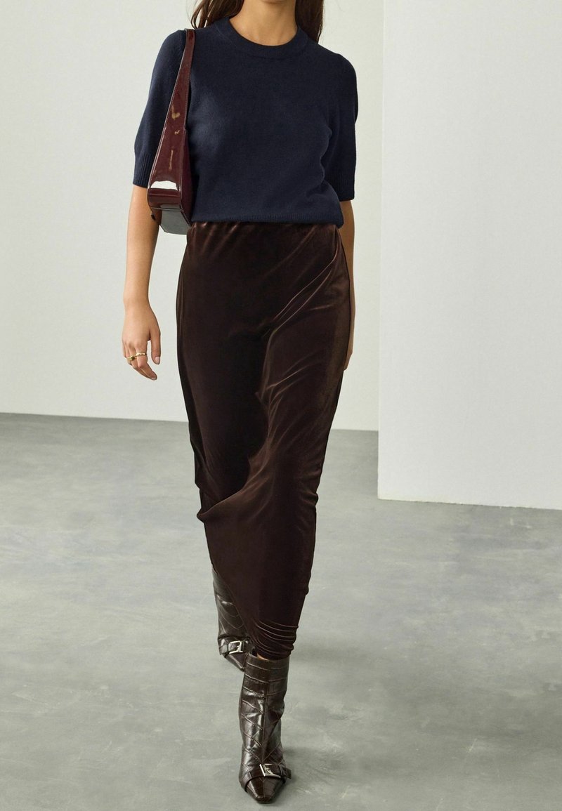 Femme portant un pull manches courtes bleu marine, une jupe en velours marron foncé, des bottes en cuir marron pointues et tenant un sac à bandoulière grenat.