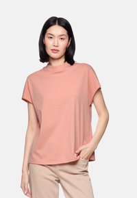 Peach-colored short-sleeve T-shirt with a high neckline, featuring metallic dot embellishments and a relaxed fit, paired with beige pants.