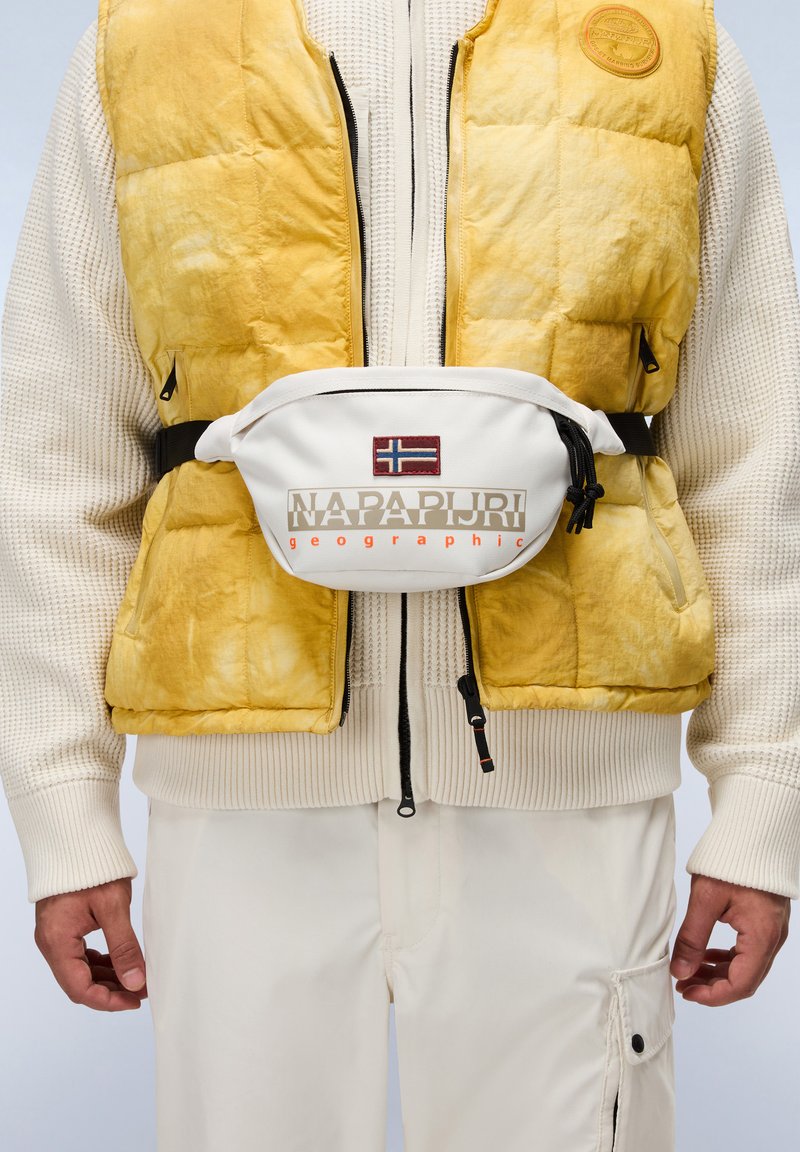 Gilet giallo gonfiato sopra un maglione lavorato a maglia bianco, abbinato a una borsa a tracolla bianca con un logo a bandiera rossa e blu e il testo "NAPAPIJRI".