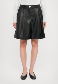 Sorte falske skinnshorts med plisserte detaljer, med frontknapp og A-formet silhuett, kombinert med svarte flate sko.