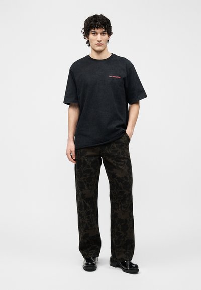 Νεαρός με σγουρά σκούρα μαλλιά, ντυμένος με μαύρο κοντομάνικο μπλουζάκι και σκούρο παντελόνι με λουλουδάτο μοτίβο, στέκεται μπροστά σε απλό φόντο.