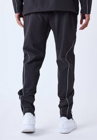 Pantalones deportivos negros con ribetes blancos, que presentan un corte relajado, cinturilla elástica y tobillos ajustados. Combinados con zapatillas blancas.