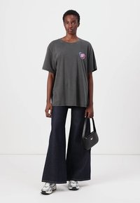 T-shirt oversize grigia con una graphic a forma di cuore sul davanti, abbinata a jeans a zampa d'elefante scuri e sneakers argento. Una semplice borsa nera completa il look.