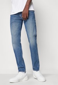 Μπλε τζιν παντελόνι με άνετη εφαρμογή, ξεθωριασμένη υφή και διακριτές ραφές. Συνδυασμένα με λευκά αθλητικά παπούτσια. Απλή σχεδίαση.