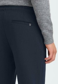 Temno modre hlače iz teksturiranega materiala, z zadnjim žepom in majhnim srebrnim logotipom. Pas je gladek in strukturiran.